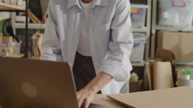 Kadın yöneticinin teslimat ofisinde çalışırken yanında karton kutuyla masada dizüstü bilgisayar kullanırken görüntüsünü yukarı kaldır.