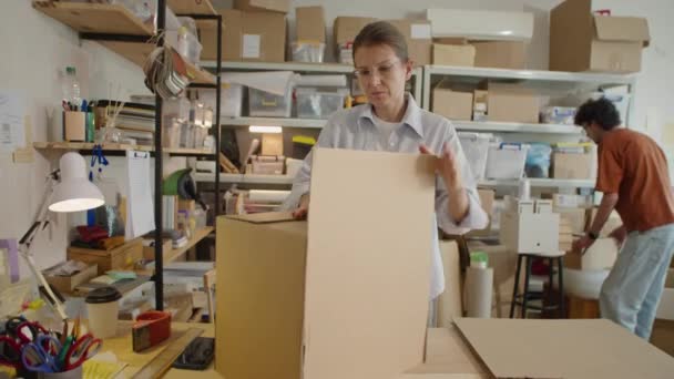 配達サービスオフィスで男性同僚と働いている間接着テープを使用し 出荷のための段ボール箱を作る女性 — ストック動画
