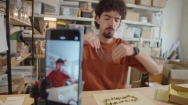 Genç adamın boncuklu kolyeler gösterirken ve kamerada konuşurken kameraya odaklan. Video bloğunu akıllı telefondan çekerken ya da atölyede canlı yayın yaparken.