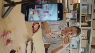 Dikey çekimde kadın el yapımı mücevherleri gösteriyor ve video reklamcılığını çekerken kameraya yansıtıyor ya da akıllı telefonlu DIY vlog 'u tripoda çekiyor.