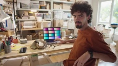 Genç erkek tasarımcının atölyede malzeme paleti ve dizüstü bilgisayarla oturduğu orta boy bir fotoğraf.