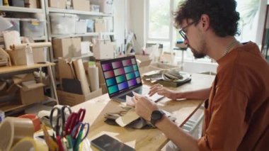 Genç erkek tasarımcı paletleri kontrol ediyor, atölyede çalışırken dizüstü bilgisayardan malzeme ve renk seçiyor