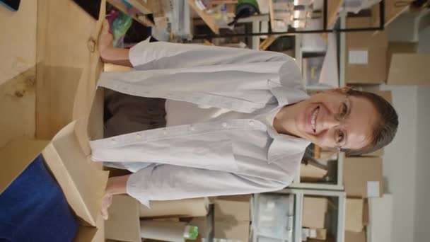 デスクの上に小包を置いて オンラインストア倉庫で仕事の日にカメラのためにポーズする笑顔の女性の垂直ショット — ストック動画