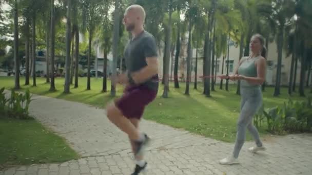 在公园进行户外运动时 穿着运动服的男子高膝跑步时 女私人教练拿着带缠在他腰间的阻力带 全速前进 — 图库视频影像
