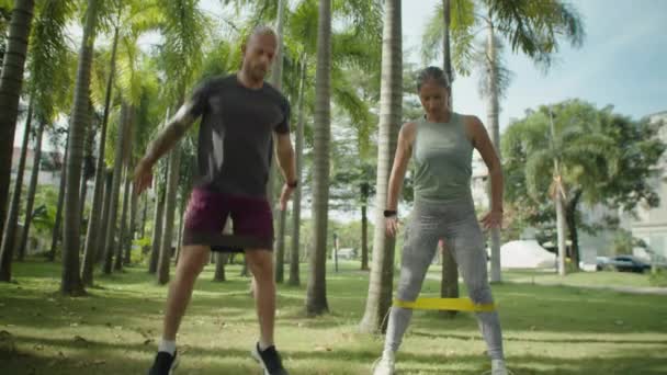 穿着运动服的运动型情侣们与阻力带一起蹲着 男人在公园的室外锻炼中同时跳着跳着 — 图库视频影像