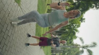Spor kıyafetli bir kadının, belinde direnç bandı tutan erkek antrenörünün yardımıyla dışarıda diz üstü yürürken dikey çekimi.