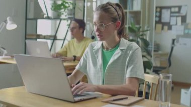 Genç bayan ofis çalışanının, mikrofonla çevrimiçi konuşma ve masaüstü bilgisayarında daktilo ile orta boy görüntüsü.