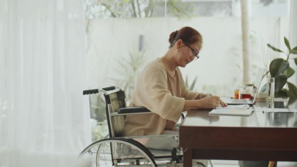 デスクで車椅子に座っている成熟したアジア女性のコピースペースを備えた中型フルショット ノートを作成し 書類を調べ 自宅で働いている間にラップトップを使用する — ストック動画