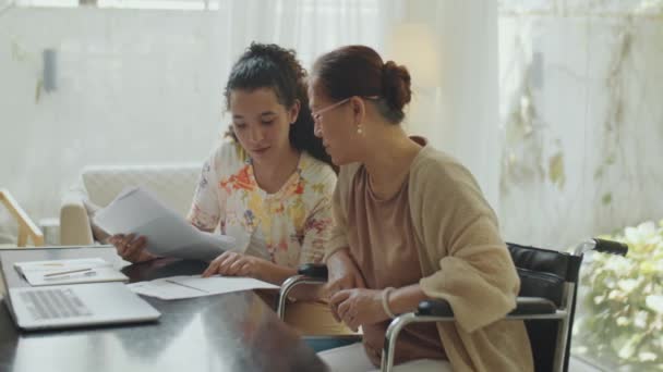 若い女性財務顧問が自宅で相談中に車椅子でアジア人女性と紙とラップトップを使用して議論 — ストック動画