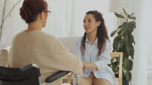 車椅子の若い女性介護者と高齢女性が手を握り 自宅でリビングルームで会話をする中型ショット — ストック動画