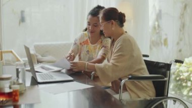 Genç bayan finans danışmanı ve tekerlekli sandalyedeki yaşlı Asyalı kadının iş belgelerini tartışırken ve ev danışmanlığı sırasında yatırım planlarken orta ölçekli bir fotoğrafı.