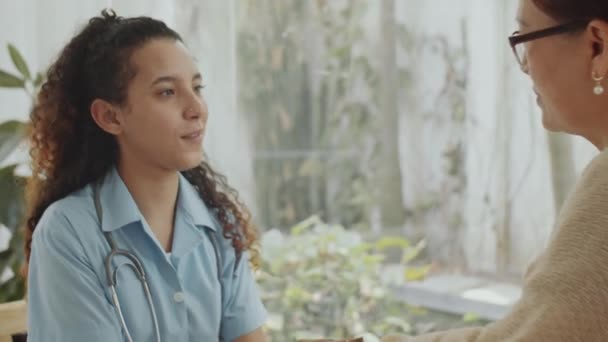医用制服の若い女性看護師が手を握り 自宅で彼女を訪問しながら車椅子で年配の女性と会話する — ストック動画