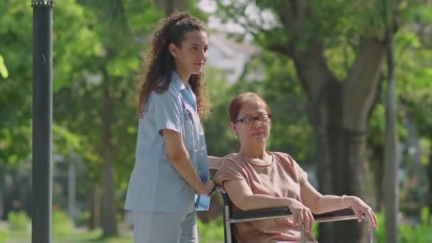 若い女性介護士が車椅子で陽気なアジア人女性とチャットしながら 夏の日に屋外で一緒に時間を過ごす中長いショット — ストック動画