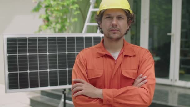 身着橙色工作服 头戴硬礼帽 头戴双臂 自信的男性技师站在屋前太阳能电池板旁边的中照照片 — 图库视频影像