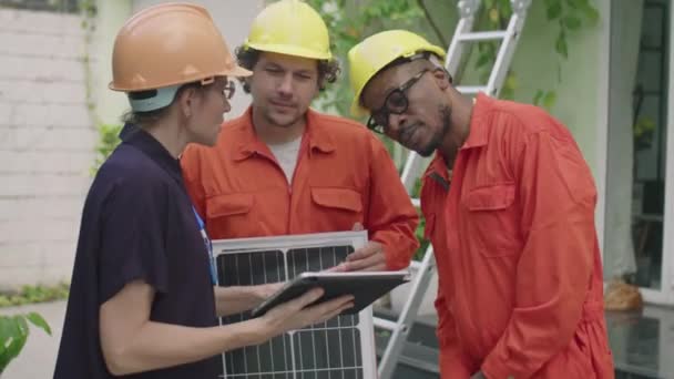 戴硬帽的女工程师使用数字平板电脑 并指导多族裔的光电池安装人员穿着工作服站在屋前的太阳能电池板上 — 图库视频影像