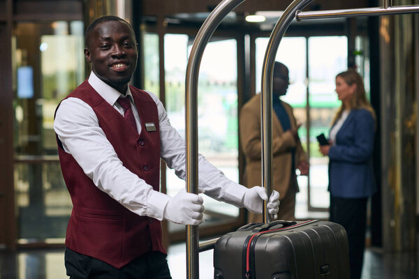 Portrait of African American doorman smiling at camera while carrying trolley with baggage