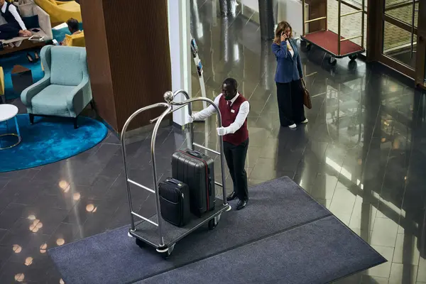stock image High angle view of African American doorman carrying the trolley with luggage in lobby of the hotel