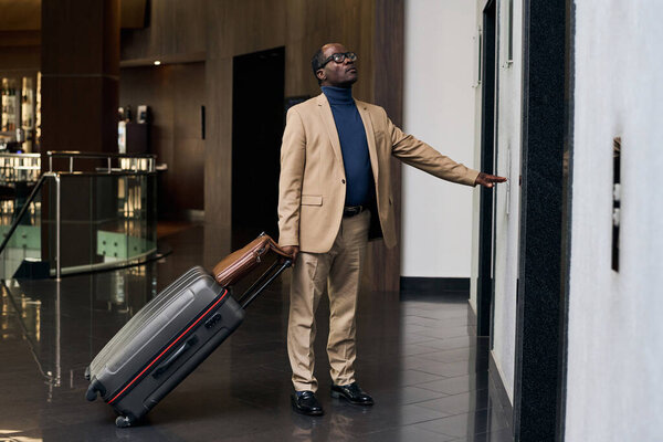 African American man with luggage standing in hall of the hotel and pushing the button of elevator