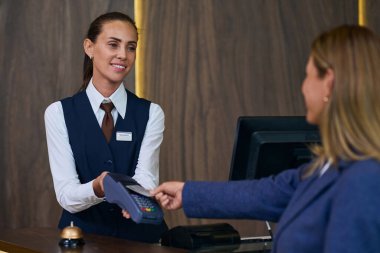 Genç resepsiyonist otelde kayıt yaptırırken müşteriye ödeme için terminal veriyor.