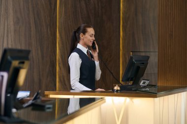Üniformalı bir yönetici tezgahın arkasında durup bilgisayarla uğraşırken telefon başında oda ayırtıyor.
