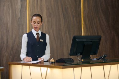 Üniformalı ciddi bir resepsiyonist oteldeki resepsiyon masasının arkasında belgeleri imzalıyor.
