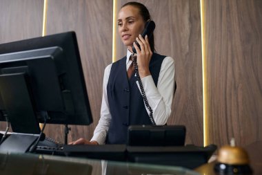 Üniformalı bir yönetici bilgisayarla tezgahın arkasında duruyor ve konuğu telefonun yanına kaydediyor.