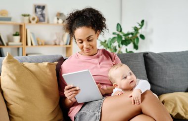 Genç anne oturma odasında kanepede oturup bebeğiyle oynarken tablet bilgisayarda video izliyor.