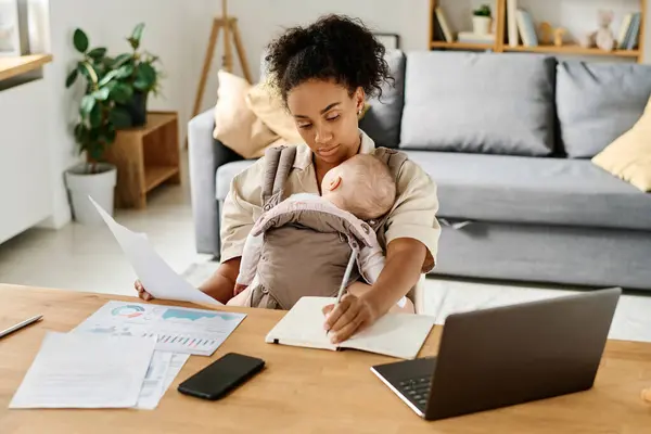 Young woman working working with documents and computer at her workplace at home with her baby sleeping in sling