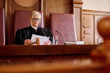 Deneyimli siyah önlüklü kadın yargıç duruşma sırasında mikrofonun önünde otururken hukuki kağıt belgeler okuyor.