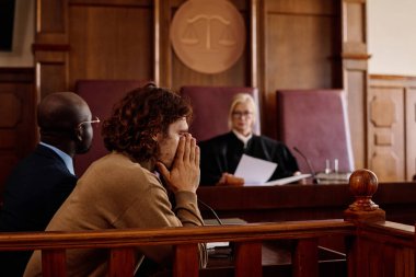 Genç endişeli erkek şüpheli, yargılama sırasında yargıcın karar vermesine karşı kameranın önünde otururken elleriyle yüzünü kapatıyor.