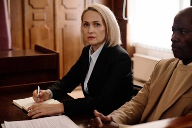 Sarışın kadın jüri veya savcı kameraya bakıyor ve not defterine not alıyor. Olgun Afrikalı Amerikalı erkek meslektaşın yanında oturuyor.