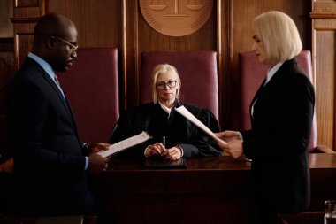 Kendine güvenen olgun bir kadın yargıç, duruşma sırasında davacı ile savunma tarafı arasında masa başında oturuyor.