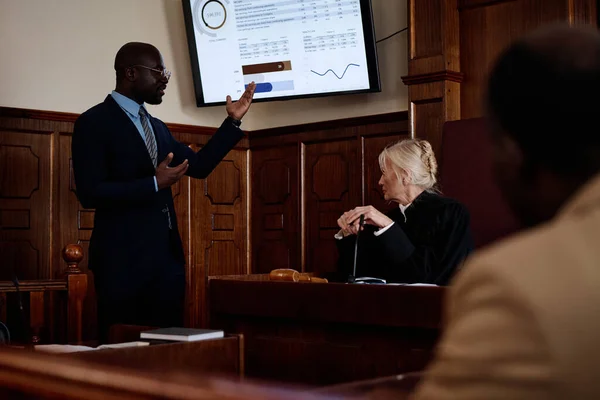 Confident African American lawyer in formalwear pointing at screen with summary of results while explaining data to mature female judge