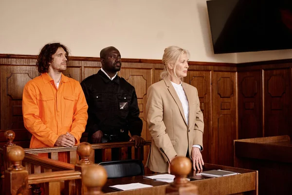 African American policeman standing between young male prisoner in cuffs and orange jumpsuit and mature female attorney during trial session