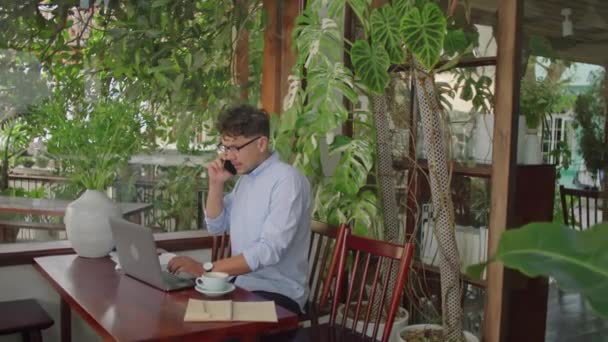 トロピカルプラントで屋外カフェに座り 携帯電話で話したり リモートジョブをしながらラップトップで作業している若いビジネスマンの中長いショット — ストック動画