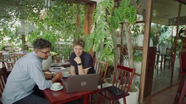ラップトップを使用して男性と女性の同僚のハイアングルショットと緑の植物で装飾された屋外カフェのビジネスプロジェクトで一緒に作業しながら議論 — ストック動画