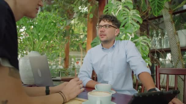 若いビジネスマンは テーブルの上にラップトップとデジタルタブレットで会議でコーヒーの上に女性の同僚と話す — ストック動画