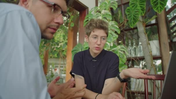 男性と女性のビジネスパートナーは ラップトップを使用して 熱帯植物で装飾された屋外カフェでのプロジェクトで一緒に作業しながら議論しています — ストック動画