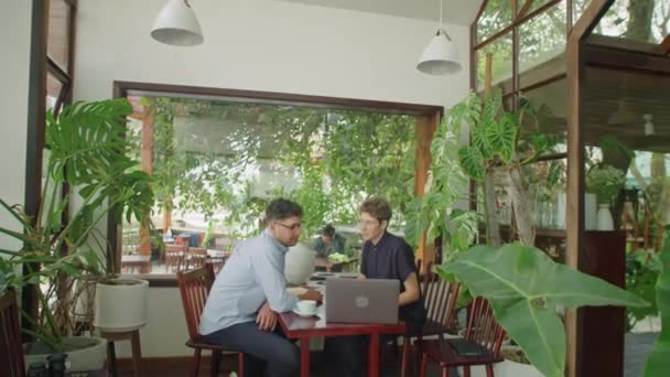 ラップトップでプロジェクトを議論し 緑の植物で装飾された屋外カフェで非公式ミーティングでコーヒーを飲むビジネスマンとビジネスマンのショットでズーム — ストック動画
