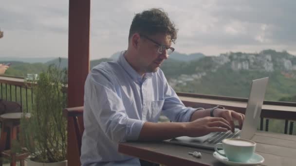 在山顶的咖啡店平台上工作时 商人在笔记本电脑上打字 桌上放着一杯卡布奇诺咖啡的中景照片 — 图库视频影像
