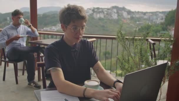 年轻女商人在山顶咖啡店的阳台上远程工作时 一边用笔记本电脑一边记笔记 一边拍中期照片 男人在后台喝咖啡 — 图库视频影像