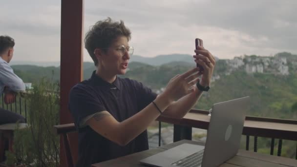 年轻女子一边在山景咖啡馆的阳台上与笔记本电脑一起工作 一边用手机通过视频通话聊天 — 图库视频影像
