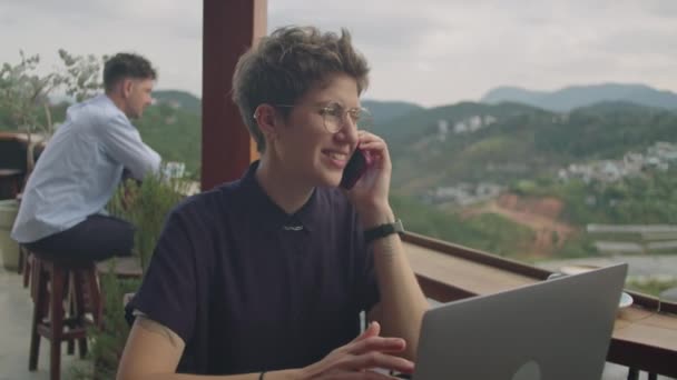 年轻的女商人一边用手机聊天 一边用笔记本电脑在山景尽收眼底的咖啡馆平台上工作 — 图库视频影像