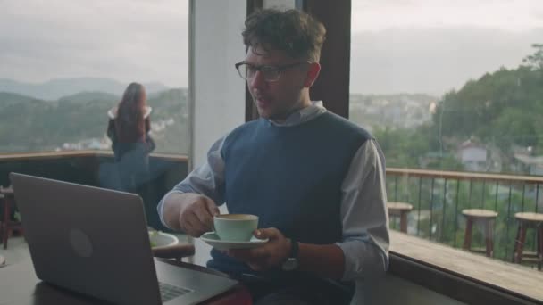 中景照片中的年轻商人喝咖啡 在可以俯瞰山景的全景窗口咖啡店工作 — 图库视频影像