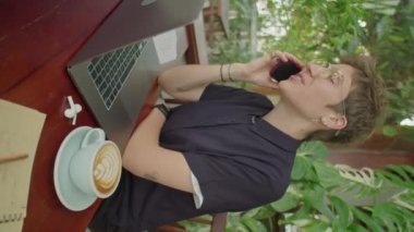 Kapuçino ve laptopla açık kafede oturan ve cep telefonuyla konuşan kısa saçlı genç iş kadınının dikey görüntüsü.