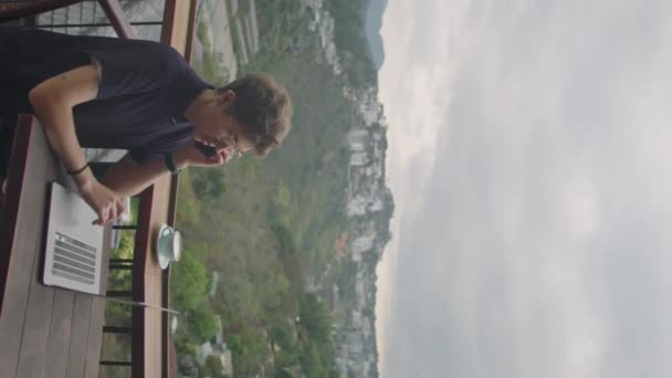風光明媚な山の景色を望むカフェテラスでラップトップで遠隔操作しながら携帯電話で通話する若いビジネスマンの垂直ショット — ストック動画