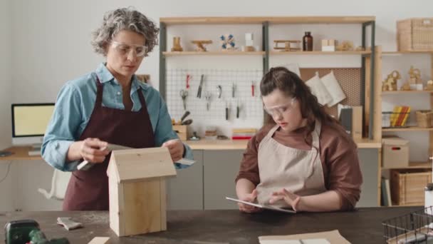 エプロンのダウン症候群とデジタルタブレットを使用して保護眼鏡を持つ女性の同僚は 大工のワークショップで昼間の定規で木製の鳥小屋を測定 — ストック動画