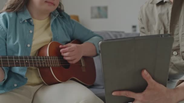 ソファーに座り ギターを弾き 家庭での音楽レッスン中に女性教師が保有するデジタルタブレットのスクリーンを見る少女のミディアムクローズアップショット — ストック動画