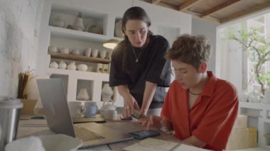 Çömlekçilik atölyesinde iş günlerinde bilgisayar ve akıllı telefon kullanan iki bayan meslektaşın orta boy görüntüsü.