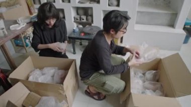 Genç Asyalı çömlekçilerin el yapımı seramiklerini müşteriye göndermek için karton kutulara paketledikleri yüksek açılı bir fotoğraf.